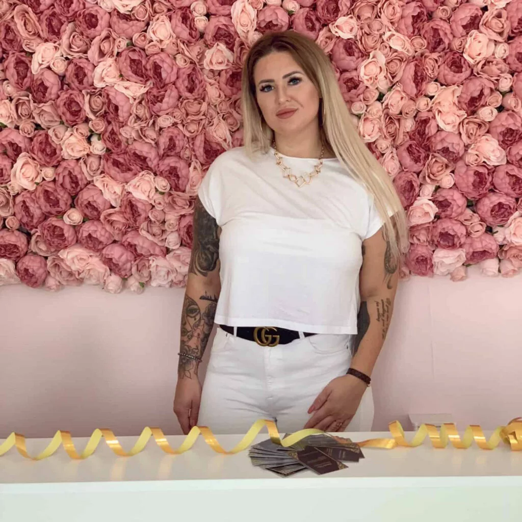 Eine Frau mit langen blonden Haaren steht vor einer mit rosa Rosen geschmückten Wand. Sie trägt ein weißes Oberteil und eine schwarze Hose mit einem dekorativen Gürtel. Eine goldene Halskette schmückt ihren Hals. Auf dem Tisch vor ihr liegen ein gelbes Band und Karten.