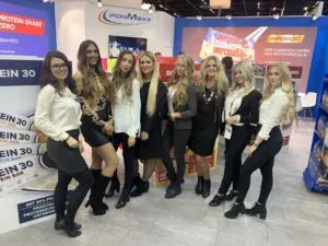 Eine Gruppe von acht Frauen posiert gemeinsam an einem Messestand. Sie stehen auf einem gefliesten Boden und tragen eine Mischung aus legerer und formeller Kleidung. Im Hintergrund sind Werbematerialien mit Logos und Produktbildern zu sehen.