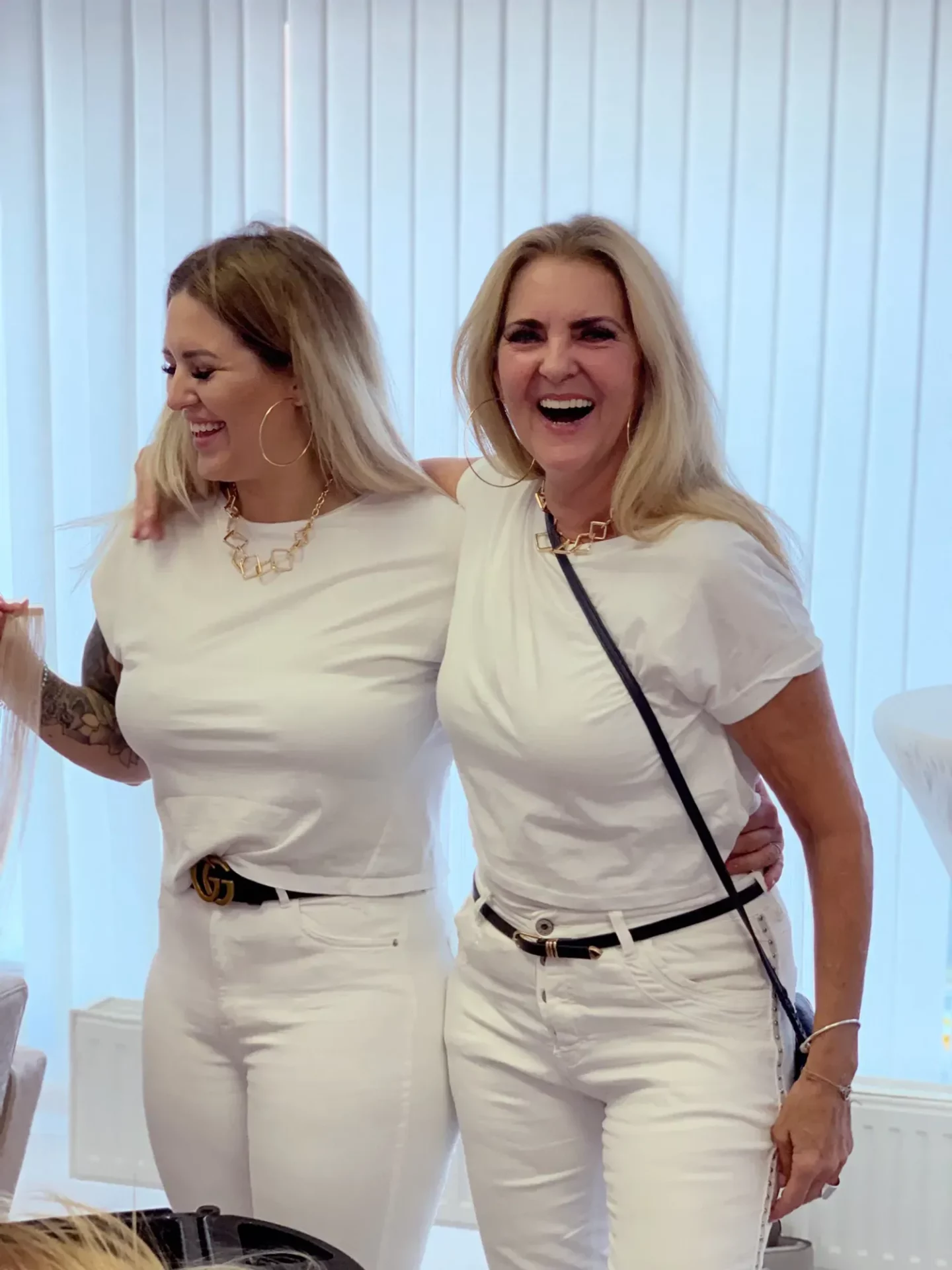 Zwei Frauen stehen zusammen vor vertikalen Jalousien, beide tragen weiße Oberteile und Hosen. Sie lächeln, wobei eine Frau den Arm um die andere gelegt hat. Beide Frauen tragen große Creolen und Gürtel.