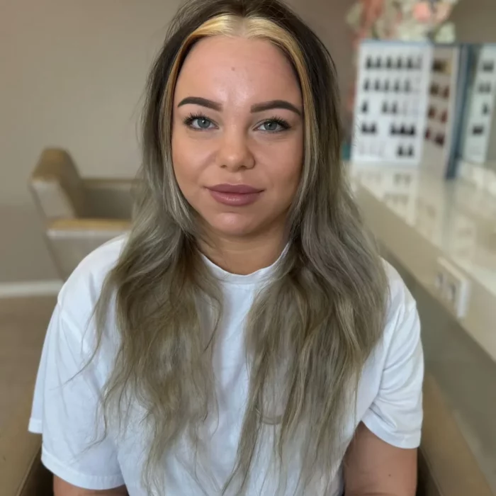 Eine Frau mit langen, welligen Haaren und einer blonden Strähne vorne sitzt in einem Friseurstuhl und trägt ein weißes T-Shirt. Im Hintergrund sind Haarfarbenproben und eine beige Wand mit Blumendekor zu sehen.