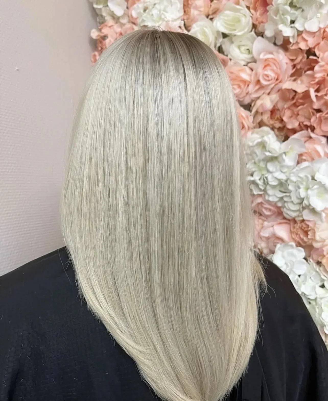 Eine Person mit langen, glatten platinblonden Haaren ist von hinten zu sehen. Im Hintergrund ist eine Wand aus pastellfarbenen Rosen und weißen Blumen zu sehen.