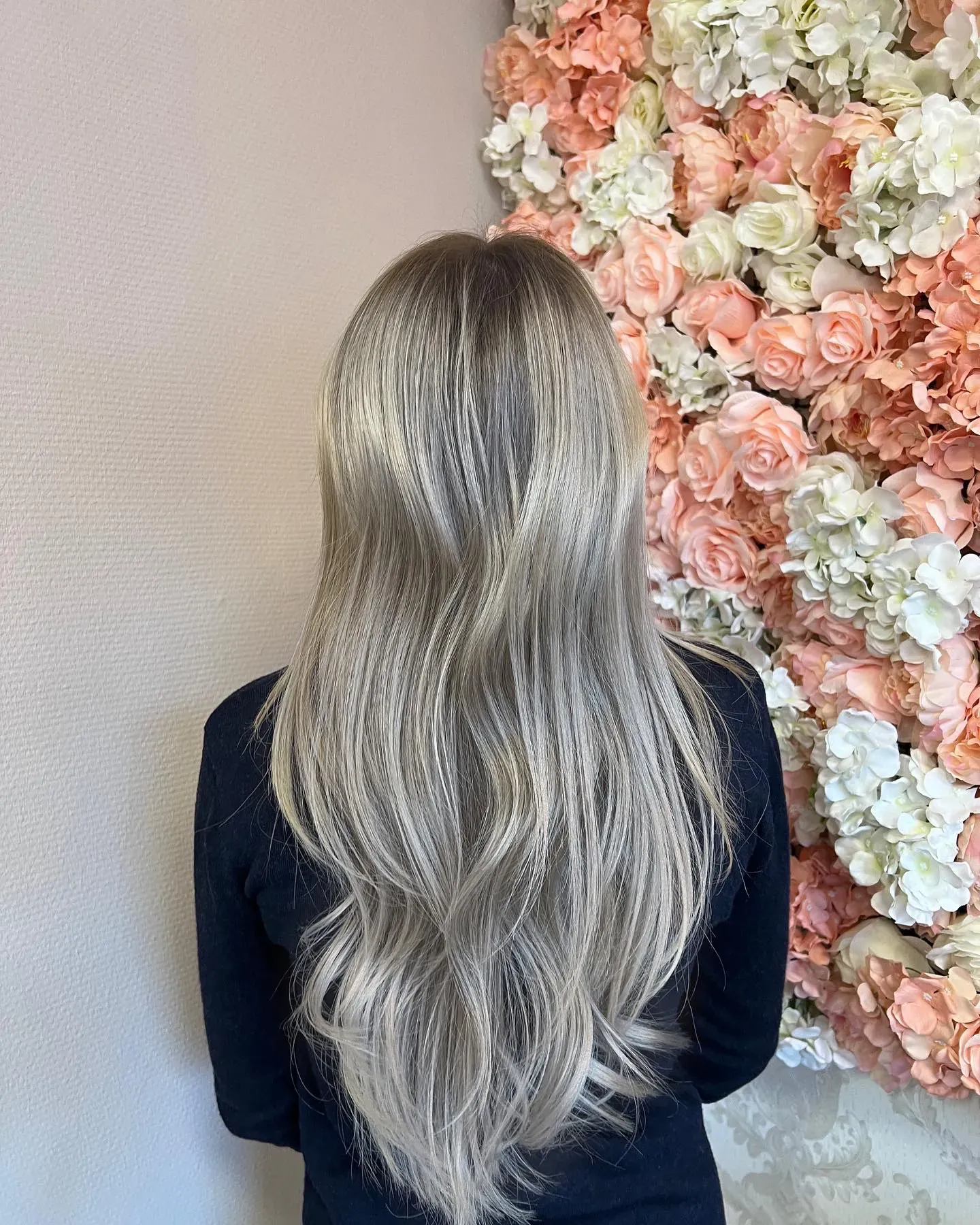 Eine Person mit langem, welligem platinblondem Haar steht mit dem Rücken zur Kamera. Sie trägt ein schwarzes Oberteil. Im Hintergrund ist eine Wand zu sehen, die mit zahlreichen künstlichen Blumen in Weiß-, Pfirsich- und Rosatönen geschmückt ist, was eine optisch ansprechende und farbenfrohe Kulisse schafft.