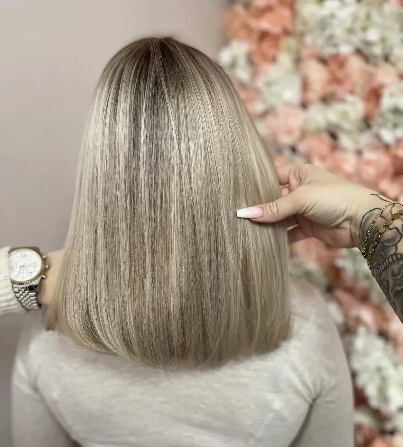 Eine Person mit glatten, schulterlangen blonden Haaren ist von hinten zu sehen. Eine Hand mit Uhr und Tattoo ordnet die Haare auf der rechten Seite. Im Hintergrund ist eine Wand mit weißen und pfirsichfarbenen Blumen zu sehen.