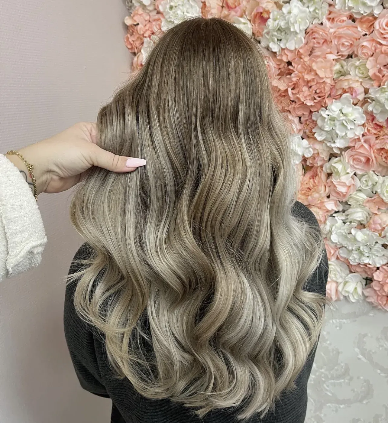 Eine Person mit langen, welligen, aschblonden Haaren ist von hinten zu sehen. Eine Hand hält sanft eine Haarsträhne. Im Hintergrund ist eine Wand mit rosa und weißen Blumendekorationen zu sehen.