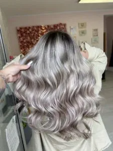 Eine Person mit langen, welligen Haaren ist von hinten zu sehen. Das Haar hat einen silbernen und aschblonden Ombre-Effekt, wobei dunklere Haarwurzeln in hellere Spitzen übergehen. Sie trägt ein beiges Oberteil und eine Hand hält eine Haarsträhne.