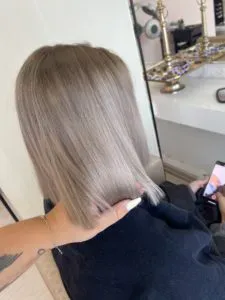 Eine Person mit kurzen, glatten, silberblonden Haaren sitzt und schaut auf ein Telefon. Eine Hand mit langen Nägeln berührt das Haar. Im Hintergrund sind ein Spiegel und ein Regal mit Gegenständen zu sehen.