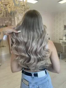 Eine Frau mit langen, welligen blonden Haaren ist von hinten zu sehen. Sie trägt ein ärmelloses Top und Jeans. Eine Hand hält eine Haarsträhne und zeigt die voluminösen Locken. Die Kulisse scheint ein Friseursalon mit Kronleuchter und gemusterter Tapete zu sein.