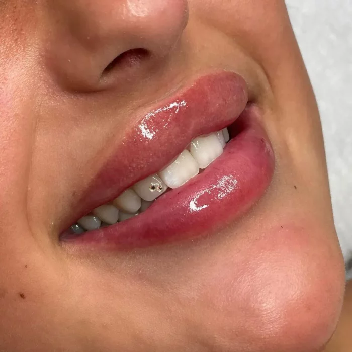 Nahaufnahme des Mundes einer Person. Man sieht glänzende, leicht rosa Lippen und weiße Zähne. Auf einem der Vorderzähne ist ein dezentes Schmuckstück zu sehen. Die Haut um den Mund herum erscheint glatt und gut beleuchtet.