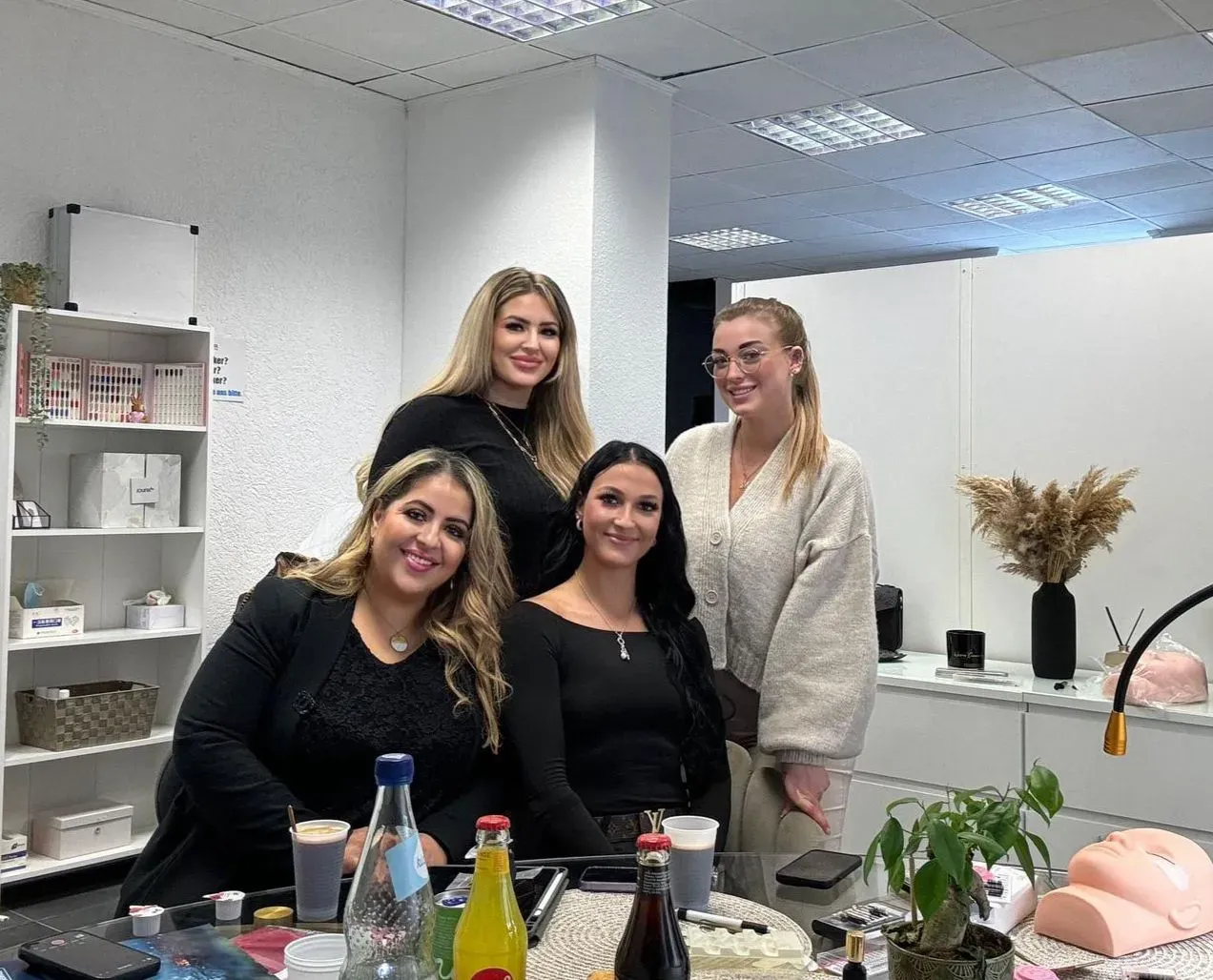 Vier Frauen sind in einem Büro versammelt, im Hintergrund stehen Regale. Zwei sitzen und zwei stehen hinter ihnen. Auf dem Tisch davor stehen Getränke und eine Topfpflanze. Sie lächeln und schauen in die Kamera.