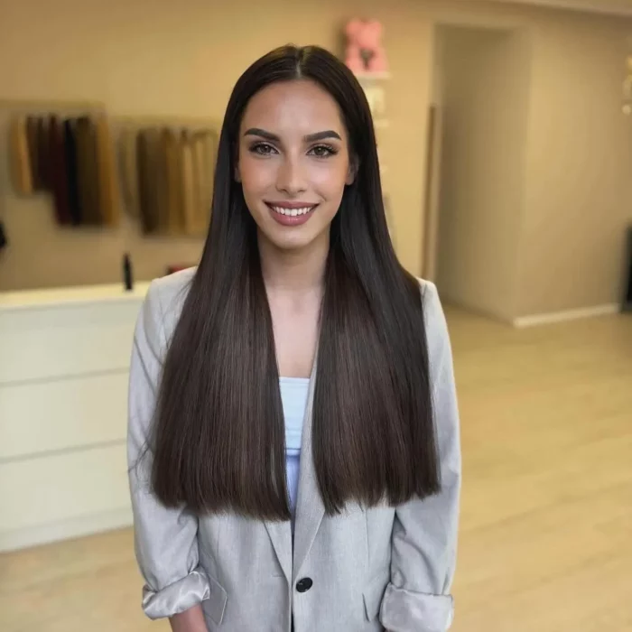 Eine Person mit langen, glatten braunen Haaren steht lächelnd im Innenbereich. Sie trägt einen hellgrauen Blazer über einem hellen Oberteil. Im Hintergrund sind ein Kleiderständer mit hängender Kleidung und eine helle Wand zu sehen.